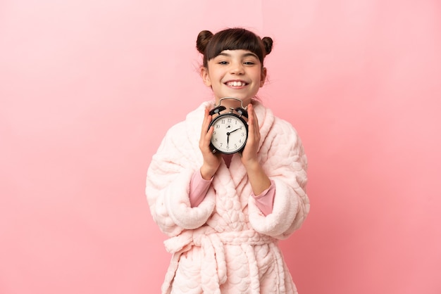 Menina caucasiana isolada na parede rosa de pijama e segurando o relógio com uma expressão feliz