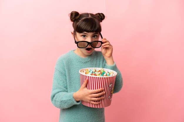 Menina caucasiana isolada na parede rosa surpresa com óculos 3D e segurando um grande balde de pipocas