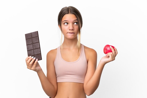 Menina caucasiana isolada no fundo branco tendo dúvidas enquanto toma um comprimido de chocolate em uma mão e uma maçã na outra