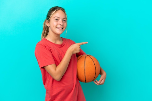 Menina caucasiana jogando basquete isolada em fundo azul, apontando o dedo para o lado