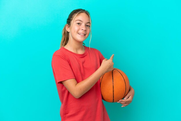 Menina caucasiana jogando basquete isolada em fundo azul, apontando para trás