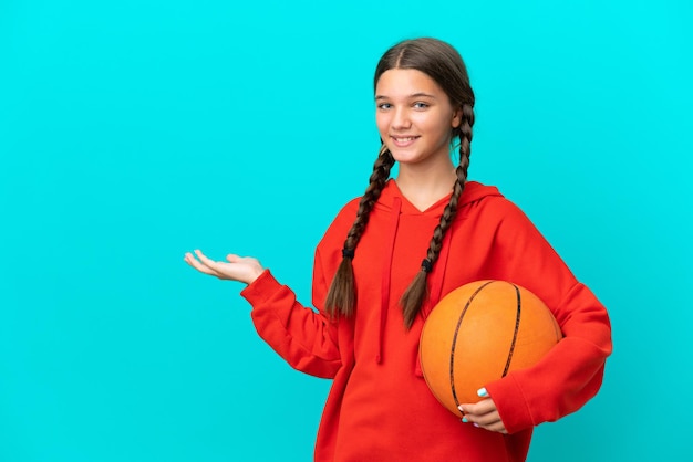 Menina caucasiana jogando basquete isolada em fundo azul, estendendo as mãos para o lado para convidar para vir