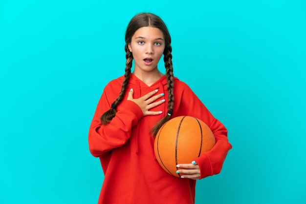 Menina caucasiana jogando basquete isolada em fundo azul surpresa e chocada ao olhar para a direita