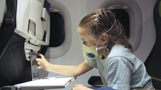 Menina caucasiana no avião com máscara protetora Criança bebê turista em aeronave com respirador jogando no smartphone móvel Covid19