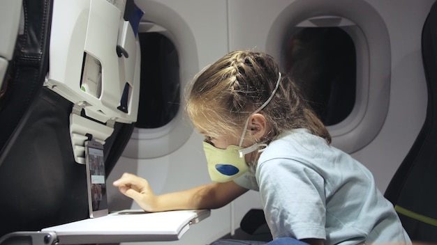 Menina caucasiana no avião com máscara protetora Criança bebê turista em aeronave com respirador jogando no smartphone móvel Covid19