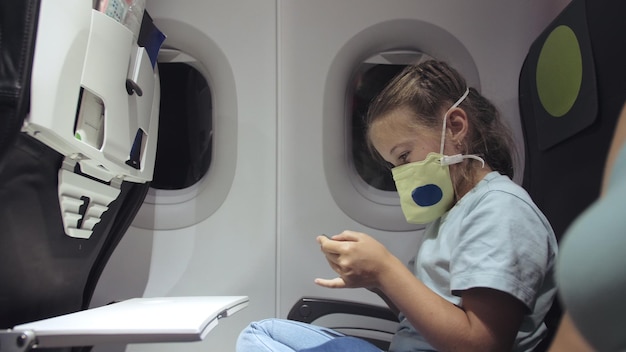 Menina caucasiana no avião com máscara protetora Criança bebê turista em aeronave com respirador jogando no smartphone móvel Covid19