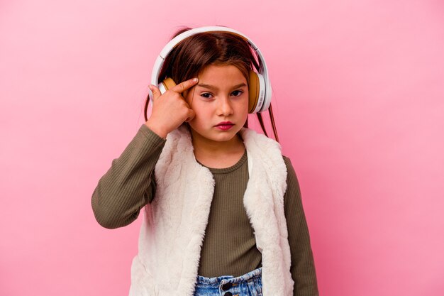 Menina caucasiana ouvindo música isolada em fundo rosa, mostrando um gesto de decepção com o dedo indicador.