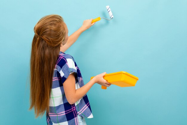 Menina caucasiana pinta uma parede com um rolo e tinta isolada em azul