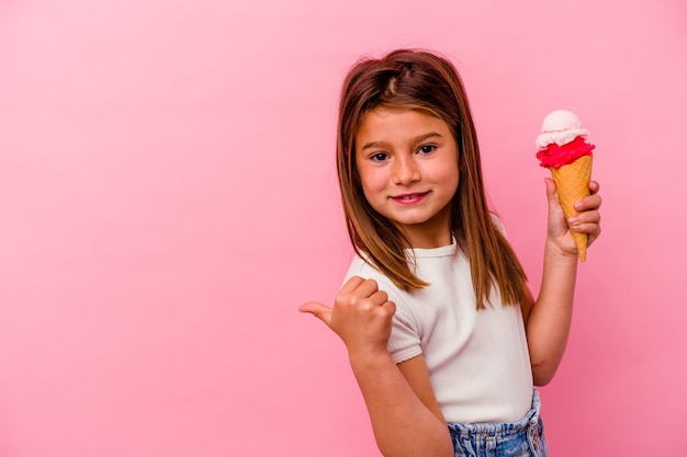 Menina caucasiana segurando sorvete isolado em pontos de fundo rosa com o dedo polegar de distância, rindo e despreocupada.