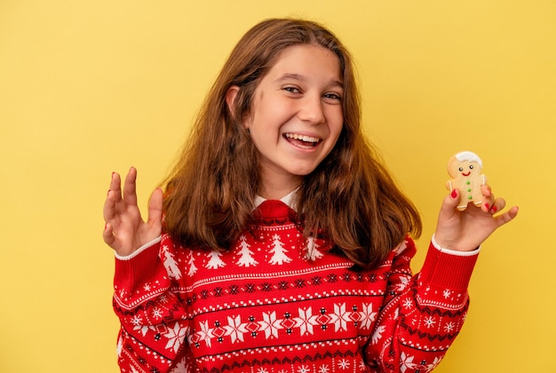 Menina caucasiana segurando um biscoito de Natal isolado no fundo amarelo, recebendo uma agradável surpresa, animada e levantando as mãos.