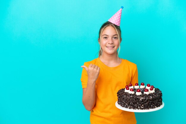 Menina caucasiana segurando um bolo de aniversário isolado em um fundo azul apontando para o lado para apresentar um produto