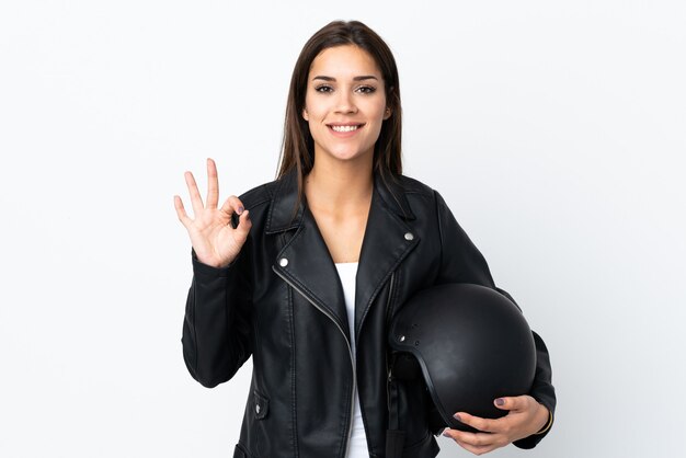 Menina caucasiana segurando um capacete de motocicleta na parede branca e mostrando um sinal de ok com os dedos