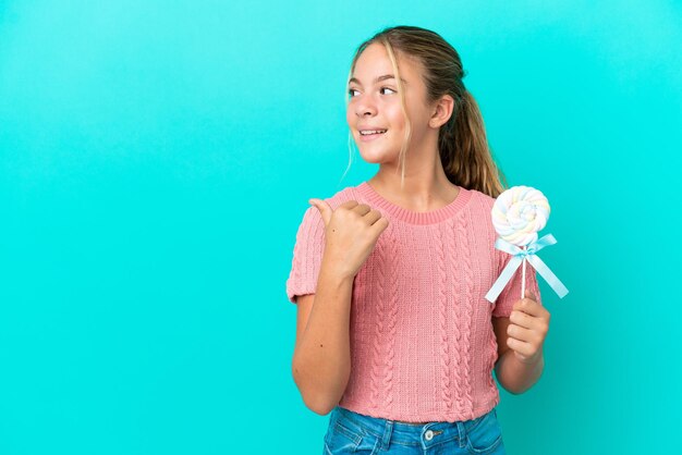 Menina caucasiana segurando um pirulito isolado em fundo azul apontando para o lado para apresentar um produto