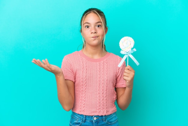 Menina caucasiana segurando um pirulito isolado em fundo azul com dúvidas ao levantar as mãos