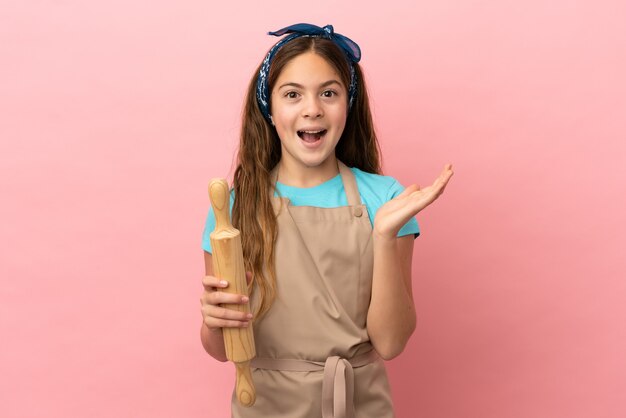 Menina caucasiana segurando um rolo de massa isolado em um fundo rosa com expressão facial chocada