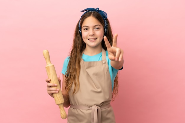 Foto menina caucasiana segurando um rolo de massa isolado no fundo rosa, sorrindo e mostrando o sinal da vitória