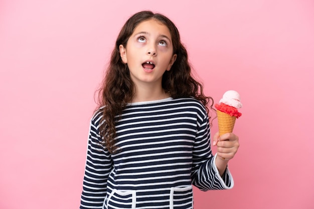 Menina caucasiana segurando um sorvete isolado no fundo rosa olhando para cima e com expressão de surpresa