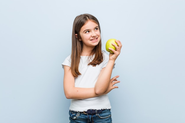 Menina caucasiana segurando uma maçã verde sorrindo confiante com braços cruzados