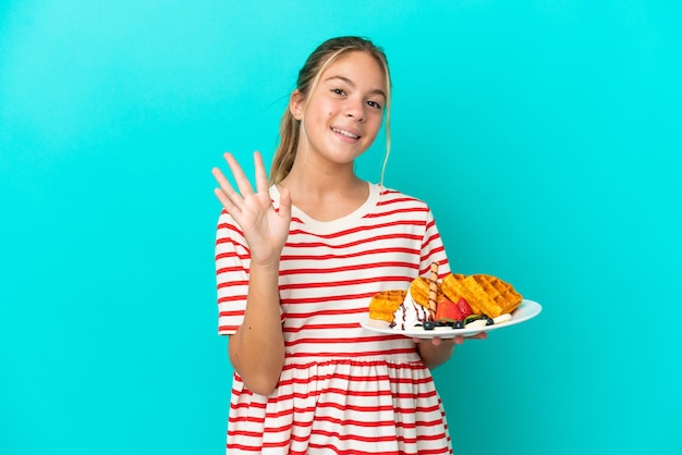 Foto menina caucasiana segurando waffles isolados em fundo azul saudando com a mão com expressão feliz