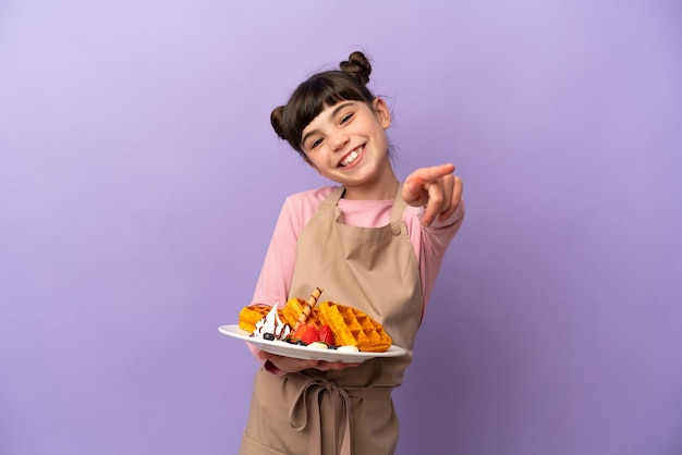 Menina caucasiana segurando waffles isolados na parede roxa apontando para a frente com uma expressão feliz