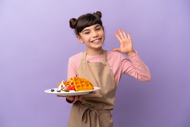 Menina caucasiana segurando waffles isolados na parede roxa saudando com a mão e expressão feliz