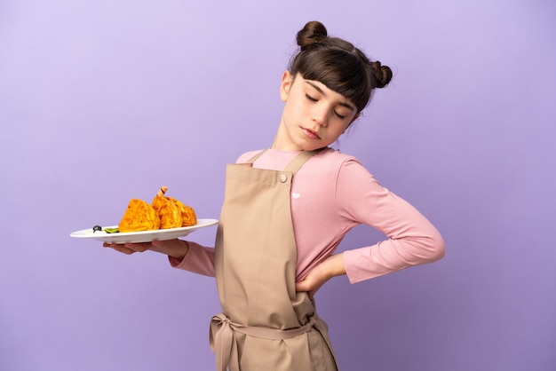 Menina caucasiana segurando waffles isolados na parede roxa, sofrendo de dor nas costas por ter feito um esforço