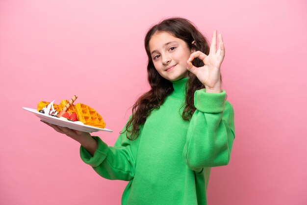 Menina caucasiana segurando waffles isolados no fundo rosa, mostrando sinal de ok com os dedos