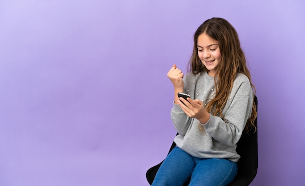 Menina caucasiana sentada em uma cadeira isolada em um fundo roxo com o telefone em posição de vitória