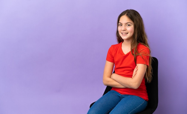 Menina caucasiana sentada em uma cadeira isolada em um fundo roxo com os braços cruzados e olhando para a frente