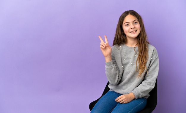 Menina caucasiana sentada em uma cadeira isolada em um fundo roxo sorrindo e mostrando sinal de vitória