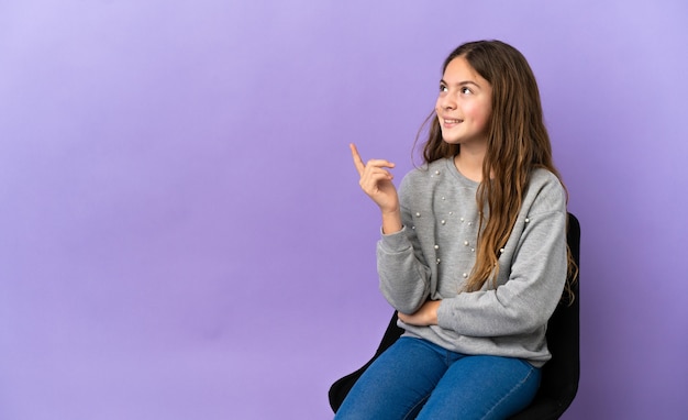 Menina caucasiana sentada em uma cadeira isolada no fundo roxo apontando para uma ótima ideia