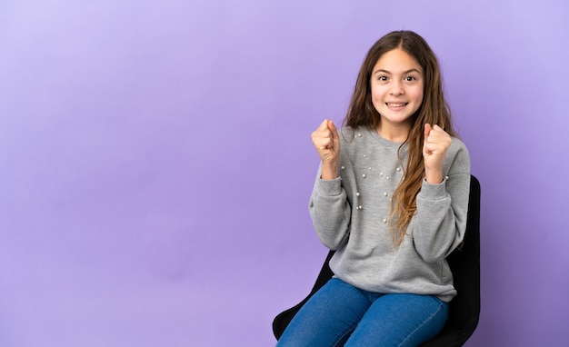 Foto menina caucasiana sentada em uma cadeira isolada no fundo roxo comemorando a vitória na posição vencedora