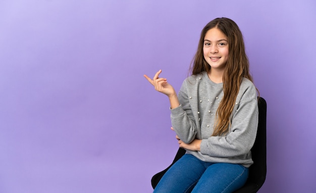 Menina caucasiana sentada em uma cadeira isolada no fundo roxo feliz e apontando para cima