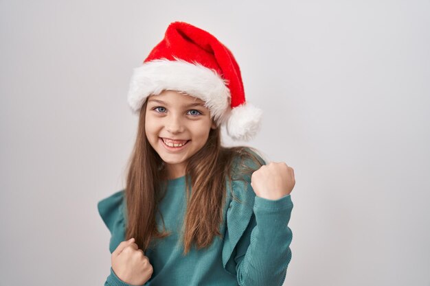 Menina caucasiana usando chapéu de natal comemorando surpresa e admiração pelo sucesso com os braços levantados e os olhos fechados