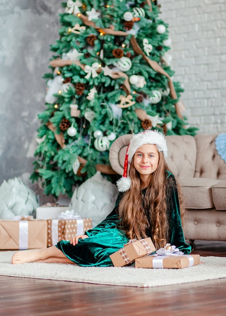 Menina chapéu de Papai Noel com presentes debaixo da árvore de Natal, sentado junto à lareira, descompacta presentes.