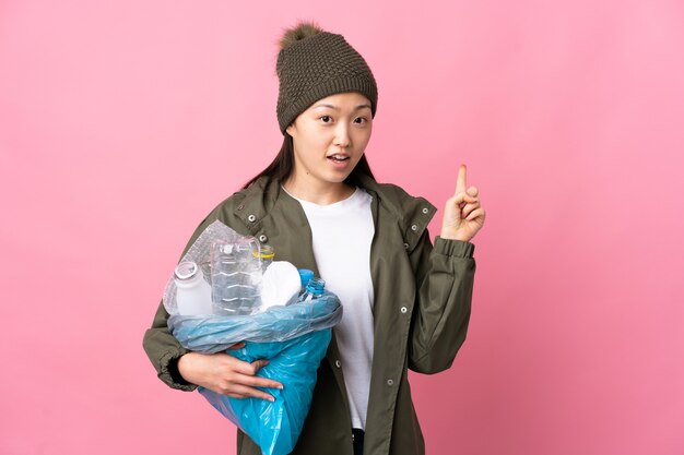 Menina chinesa segurando uma sacola cheia de garrafas plásticas para reciclar em uma rosa isolada surpresa e apontando para o lado