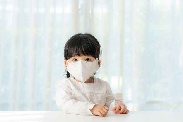 Menina chinesa vestindo máscara facial saudável, sentado na sala de estar em casa para evitar a poeira PM2.5, poluição atmosférica, poluição do ar e COVID-19.