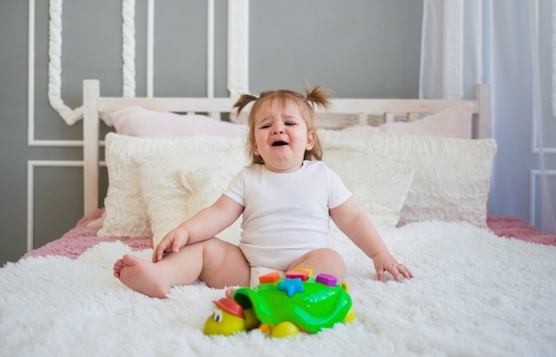 Menina chorando sentada na cama com um brinquedo classificador no quarto