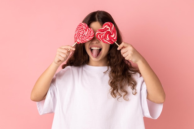 Menina cobrindo os olhos com doces em forma de coração mostrando a língua posando com expressão feliz