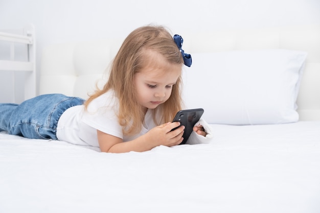 Menina com a mão engessada, deitada na cama usando o smartphone, assistindo a um desenho animado ou vídeo educacional.