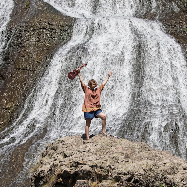 Menina com as mãos levantadas com ukulele em pé sobre uma rocha perto da cachoeira