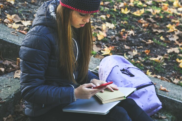 Foto menina com boné de lã sentada no parque usando smartphone estudante adolescente fazendo a lição de casa com o telefone ao ar livre