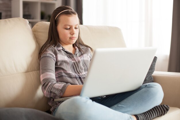 Menina com cabelo comprido, sentada no sofá da sala de estar usando o laptop.