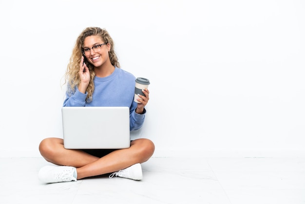 Menina com cabelo encaracolado com um laptop sentado no chão segurando café para levar e um celular