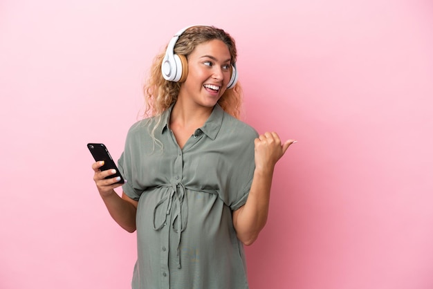 Menina com cabelo encaracolado isolado em um fundo rosa grávida e ouvindo música enquanto aponta para o lado