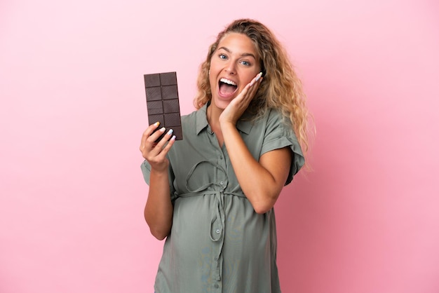 Menina com cabelo encaracolado isolado em um fundo rosa grávida e segurando um chocolate com expressão de surpresa