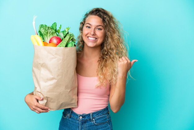 Menina com cabelo encaracolado segurando uma sacola de compras isolada em fundo verde, apontando para o lado para apresentar um produto