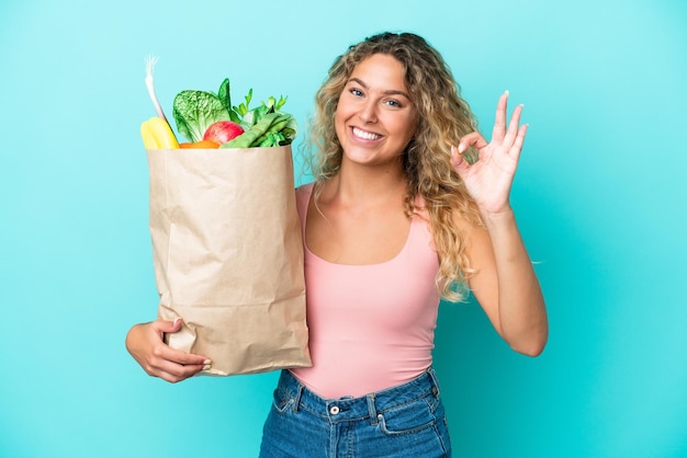 Menina com cabelo encaracolado, segurando uma sacola de compras isolada em fundo verde, mostrando sinal de ok com os dedos