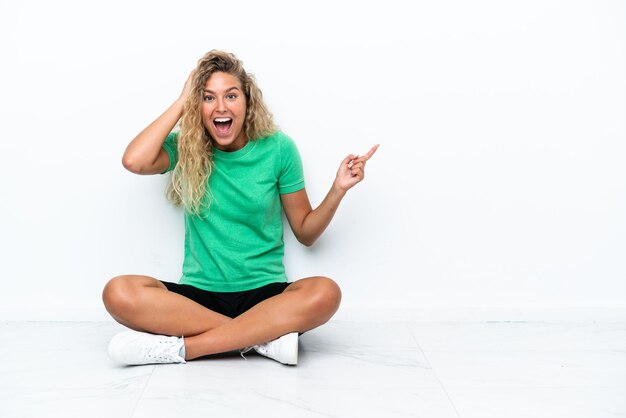 Menina com cabelo encaracolado sentada no chão surpresa e apontando o dedo para o lado