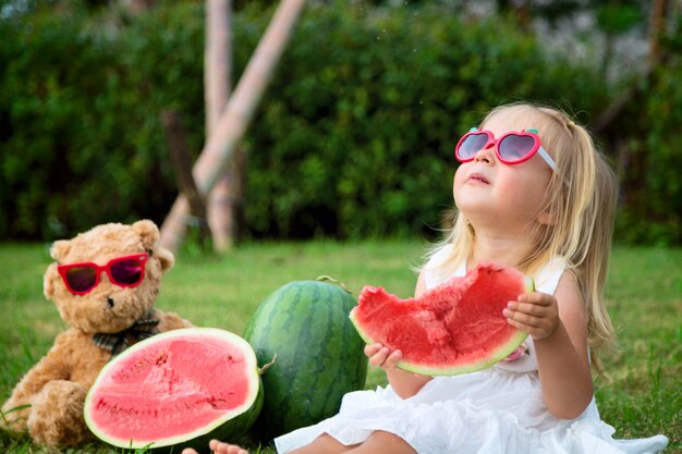 Menina com cabelo loiro em óculos de sol, comendo melancia no parque, próximo sentado urso de pelúcia